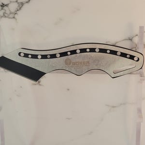 May include: A silver-colored Boker knife with a black blade. The knife has a curved design with a series of small circular cutouts along the top edge. The Boker logo is visible on the blade.