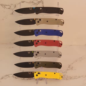 May include: A vertical display of seven folding knives with black blades and textured handles in black, tan, blue, red, gray, olive green, and yellow. Each knife features a small butterfly logo on the blade and a silver locking mechanism.