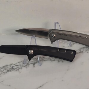 May include: Two folding knives with pointed blades and metallic handles. One knife features a matte black finish, while the other has a metallic grey finish. Both knives display the text "HARD TO HANDLE" on the handle near the pivot screw.