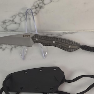 May include: A fixed-blade knife with a silver-toned pointed blade marked with the text CRKT. The handle features a textured grey and black pattern with three silver rivets. A black braided cord lanyard is attached to the end of the handle.