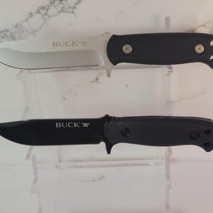 May include: Two knives with black handles and silver blades are displayed. The top knife has a silver blade with the word "BUCK" printed on it. The bottom knife has a black blade with the word "BUCK" printed on it.