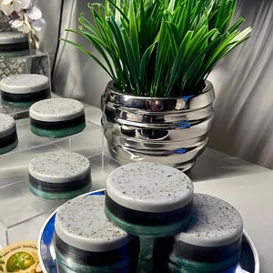 May include: Round, layered soaps with gray, black, and teal bands are stacked on a mirrored surface. The soaps are labeled "Oatmilk Soap" and "100% Vegan Handcrafted". A silver pot with green grass is in the background.