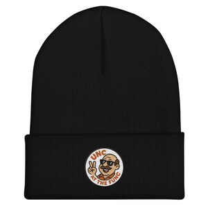 Może przedstawiać: Czarna czapka beanie z okrągłą naszywką przedstawiającą kreskówkowego mężczyznę pokazującego znak pokoju. Na naszywce widnieje napis "UNC at the Func". Czapka wykonana jest z miękkiego, dzianinowego materiału.
