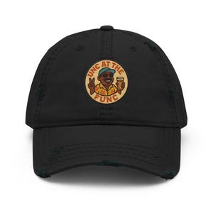 May include: Black distressed baseball cap with a curved brim. The cap features a circular patch with the text "UNC AT THE FUNC" and a cartoon illustration of a person holding a beer and giving a peace sign.