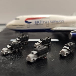 Puede incluir: Maqueta de un avión Boeing 747 de British Airways con varios camiones pequeños en blanco y negro en primer plano. El avión es blanco con detalles azules y rojos, y las palabras "BRITISH AIRWAYS" están impresas en el lateral.