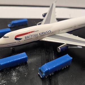 Puede incluir: Maqueta de un avión Boeing 747 de British Airways, blanco con alas grises y motores azules, con el logotipo de la aerolínea. Varios camiones de juguete azules están esparcidos alrededor del avión sobre una superficie negra.