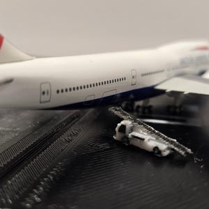 Könnte beinhalten: Ein weiß-blaues Modellflugzeug mit dem British Airways-Logo und einer kleinen Servicetreppe. Das Modell steht auf einer schwarzen Oberfläche. Das Flugzeug ist eine Boeing 747.