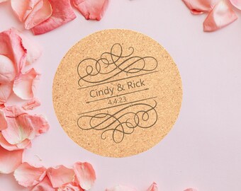 May include: A round cork coaster with a brown background and a decorative design in a dark brown color. The design includes a swirl pattern and the text "Cindy & Rick 4.4.23". The coaster is surrounded by pink rose petals.