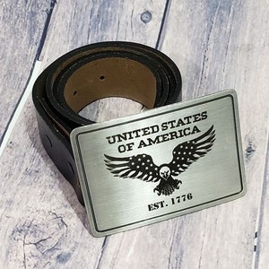 Puede incluir: Una hebilla de cinturón de plata con un águila y el texto "UNITED STATES OF AMERICA EST. 1776". La hebilla está unida a un cinturón de cuero negro.