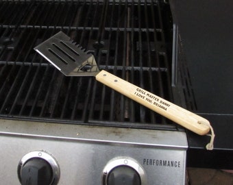 May include: A wooden spatula with a metal blade and the text "Grill Master Danny I Love You, Brianna" engraved on the handle. The spatula is resting on a black grill with three knobs.