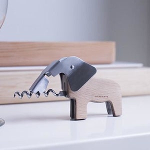 Puede incluir: Un sacacorchos de madera con forma de elefante con un sacacorchos de metal plateado y una palanca de metal plateado. El sacacorchos est&aacute; sobre una superficie blanca.