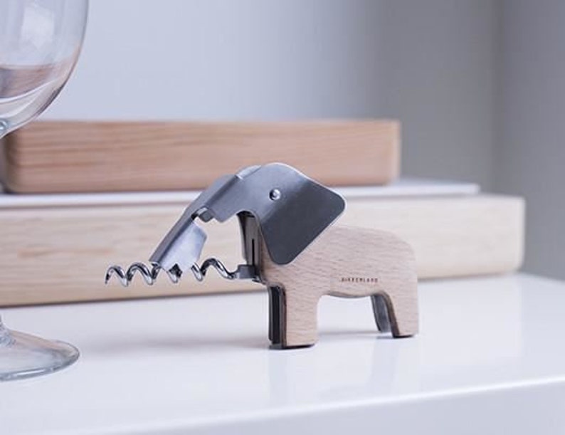 Puede incluir: Un sacacorchos de madera con forma de elefante con un sacacorchos de metal plateado y una palanca de metal plateado. El sacacorchos est&aacute; sobre una superficie blanca.
