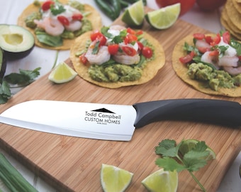 Puede incluir: Un cuchillo de chef de cerámica blanca con mango negro, descansando sobre una tabla de cortar de madera. El cuchillo tiene el texto "Todd Campbell CUSTOM HOMES" impreso en la hoja. La tabla de cortar está rodeada de limas, aguacates y cilantro.