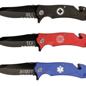 Puede incluir: Tres navajas plegables con hojas negras y mangos de colores. La navaja superior es negra con "BILL" grabado en la hoja y un símbolo de cruz blanca. La navaja del medio es roja con "TED" y un emblema de bomberos. La navaja inferior es azul con "JOHNNY" y una estrella de ambulancia.