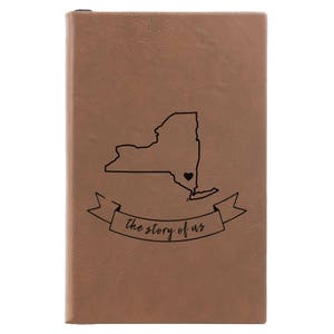 Peut inclure: Un journal en cuir marron avec un contour noir de l'&eacute;tat de New York et un petit c&oelig;ur au centre. Le journal a une banni&egrave;re qui dit "the story of us".