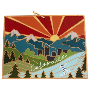 Pode incluir: Um ornamento de madeira que representa um pôr do sol estilizado sobre uma cordilheira com um horizonte urbano e um esquiador em primeiro plano. O texto "Colorado" está escrito em itálico branco sobre a imagem.