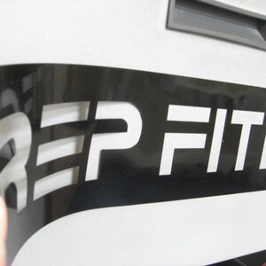 Puede incluir: Una plantilla de vinilo negro con la palabra "REP FITNESS" recortada en blanco. La plantilla se est&aacute; despegando para revelar las letras blancas.