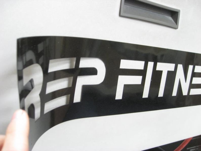 Puede incluir: Una plantilla de vinilo negro con la palabra "REP FITNESS" recortada en blanco. La plantilla se est&aacute; despegando para revelar las letras blancas.