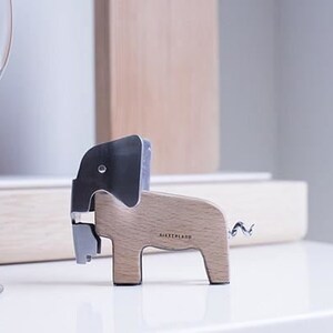 Puede incluir: Un sacacorchos de madera con forma de elefante con una cabeza de metal plateado y una cola de metal plateado. El sacacorchos est&aacute; etiquetado como "JAERTHLAND".