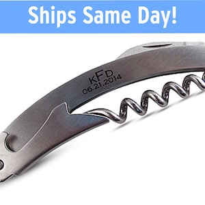 May include: A silver-colored, engraved,  wine bottle opener with a corkscrew and a bottle opener. The engraving reads "KFD 06.21.2014".