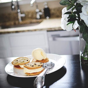 May include: Three chocolate chip cookie ice cream sandwiches on a white plate with a silver ice cream scoop. The plate is on a black surface.