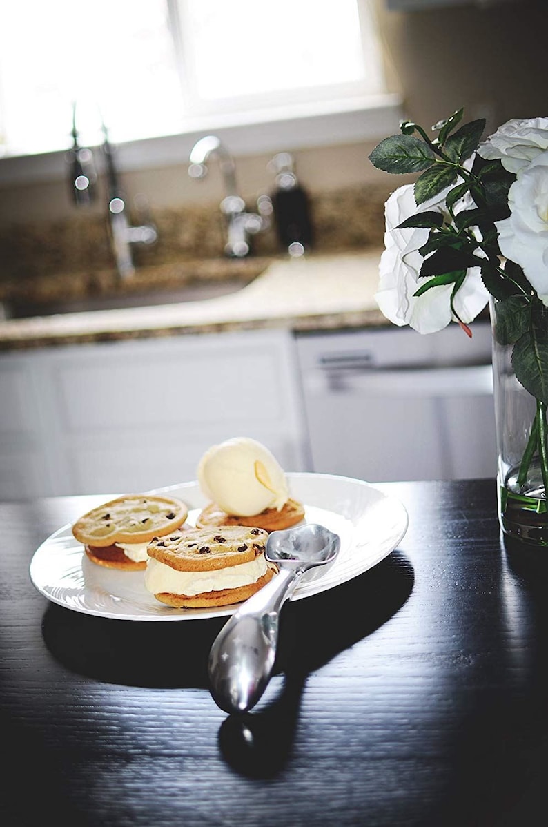 May include: Three chocolate chip cookie ice cream sandwiches on a white plate with a silver ice cream scoop. The plate is on a black surface.