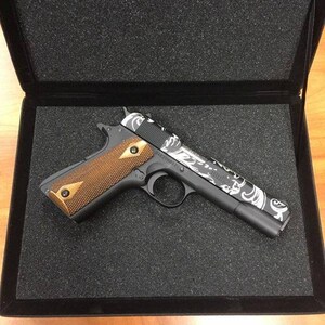 May include: A black handgun with a white and grey patterned design on the slide. The gun has a brown wooden grip and is in a black foam-lined case.