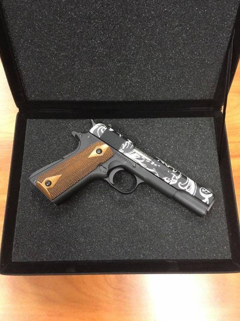 May include: A black handgun with a white and grey patterned design on the slide. The gun has a brown wooden grip and is in a black foam-lined case.