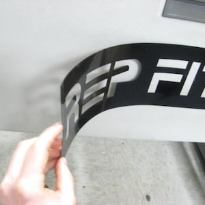 Puede incluir: Una pegatina de vinilo negro con letras blancas que dice "REP FITNESS". La pegatina se est&aacute; despegando de una superficie blanca.