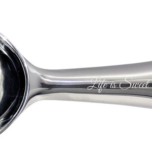 May include: A silver ice cream scoop with a curved handle and a polished finish. The handle has the words "Life is Sweet" engraved on it.