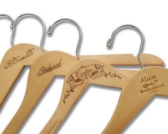 Könnte beinhalten: Vier Holzbügel mit graviertem Text. Die Bügel sind aus Naturholz mit einem silbernen Metallhaken. Der Text auf den Bügeln lautet "Bridemaid" und "Caroline Bridemaid" und "Alice Bridemaid".
