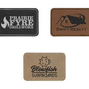 May include: Three brown leather patches with stitched edges. The first patch has a black background with a white flame and the text "Prairie Fyre Grillworx". The second patch has a brown background with a black house and the text "Swift Realty". The third patch has a brown background with a black blowfish and the text "Blowfish Surfboards".