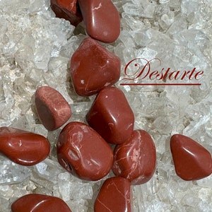 May include: A collection of ten red jasper tumbled stones on a bed of clear quartz crystals. The stones are smooth and polished, with a deep red color. The text "Destarte" is visible in the image.