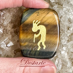 May include: A polished tiger's eye stone with a gold-colored Kokopelli design etched into the surface. The stone is a rounded rectangle shape and is being held by a hand.