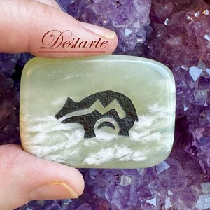 May include: A smooth, rectangular light green stone with white cloudy inclusions, featuring a black engraved bear silhouette in the center. The word Destarte is printed in a dark red script font above the stone.