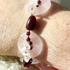 Puede incluir: Una pulsera hecha a mano con grandes discos de cuarzo rosa pálido, intercalados con cuentas de granate rojo oscuro y piedras ovaladas. Pequeños fragmentos de cuarzo blanco y detalles metálicos dorados están presentes. La palabra "Restarti" está escrita en cursiva.