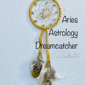 K&ouml;nnte beinhalten: Aries Astrologie Traumf&auml;nger. Dieser handgefertigte Traumf&auml;nger hat einen gelben, lederumwickelten Reifen mit einem gewebten Netz, verziert mit gr&uuml;nen und klaren Edelsteinen. Zwei gelbe Lederriemen h&auml;ngen darunter, verziert mit Federn und dekorativen Metallakzenten. Der Text "Aries Astrology Dreamcatcher" ist sichtbar.