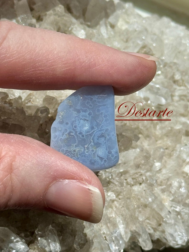 K&ouml;nnte beinhalten: Ein hellblauer Achat-Stein mit einem wei&szlig;en und grauen Muster. Der Stein wird in einer Hand vor einem Hintergrund aus wei&szlig;en Kristallen gehalten.