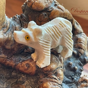 May include: A small, white, carved bear figurine with a yellow eye. The bear is standing on a piece of brown wood.