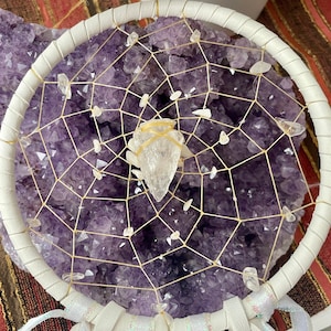 May include: A white leather-wrapped dreamcatcher with a web of beige string, adorned with clear quartz crystals. A large, pointed quartz crystal is centered, held with a beige band. The background is a cluster of purple amethyst crystals.