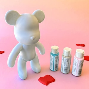 May include: A white bear-shaped figurine stands on a pink surface, next to three small paint bottles in blue, gray, and pink. Red paint splotches are scattered around the bear. The figurine is a blank canvas for customization.