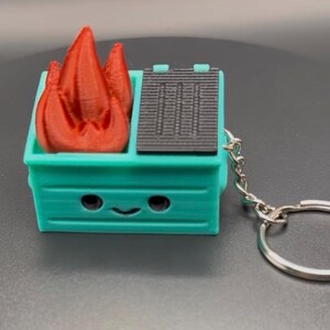 May include: A teal dumpster-shaped keychain with a smiling face, a black lid, and red flames. The keychain has a silver chain and a keyring. The dumpster is a 3D-printed novelty item.