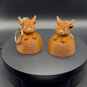 Può includere: Due portachiavi con mucche delle Highlands. Ogni portachiavi presenta una mucca marrone con una criniera bionda e piccole corna. Le mucche sono inserite in una base marrone strutturata. Ogni portachiavi ha un anello argentato.