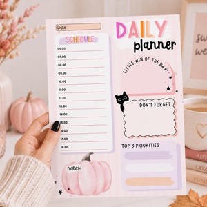 May include: A daily planner sheet with a light pink background featuring a schedule from 06:00 to 18:00, a section for little wins, a don't forget box with a black cat illustration, top three priorities, a notes section with a pink pumpkin, and stars.
