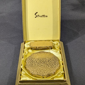 May include: A vintage Stratton compact and lipstick holder set in a gold finish, presented in a box with a satin lining. The compact has a textured, cracked-ice design. The box lid features the Stratton logo.