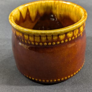May include: A small, round ceramic bowl with a brown exterior and a yellow-brown interior. The rim is yellow-brown with a dotted pattern. The bowl has a glossy finish and is likely vintage.