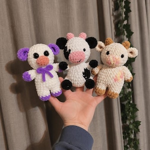 May include: Three handmade crochet animal dolls. The dolls are white with purple, black, and brown accents. One doll has purple ears and a purple bow. Another has black spots and ears. The third has brown ears and feet.