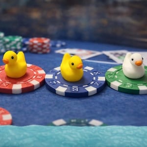 May include: Three toy ducks on poker chips. Two yellow ducks sit on red and blue chips, and a white duck rests on a green chip. Poker chips in various colors are scattered on a blue felt surface, with playing cards in the background.