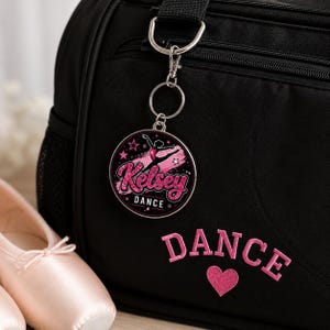 May include: A circular metal keychain featuring a pink glittery graphic of a dancer in a leap pose, the name Kelsey in pink script, and the word DANCE in white text. The keychain is attached to a black bag with the word DANCE and a heart embroidered.