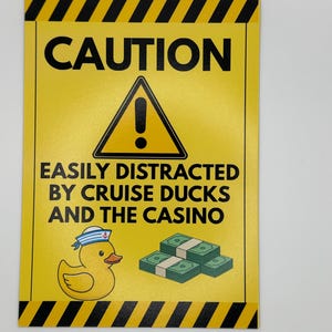 May include: A yellow sign with black diagonal stripes and the word "CAUTION" at the top. Below is a black triangle with an exclamation point, and text that reads "EASILY DISTRACTED BY CRUISE DUCKS AND THE CASINO". A cartoon duck and stacks of money are also shown.
